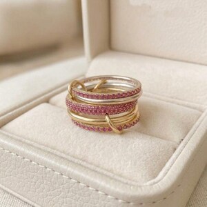 Pu&ograve; includere: Una pila di diversi anelli con fasce alternate in metallo lucido color oro e argento. Molti degli anelli sono incastonati con piccole pietre preziose rosa brillante che coprono l'intera superficie esterna delle fasce circolari.