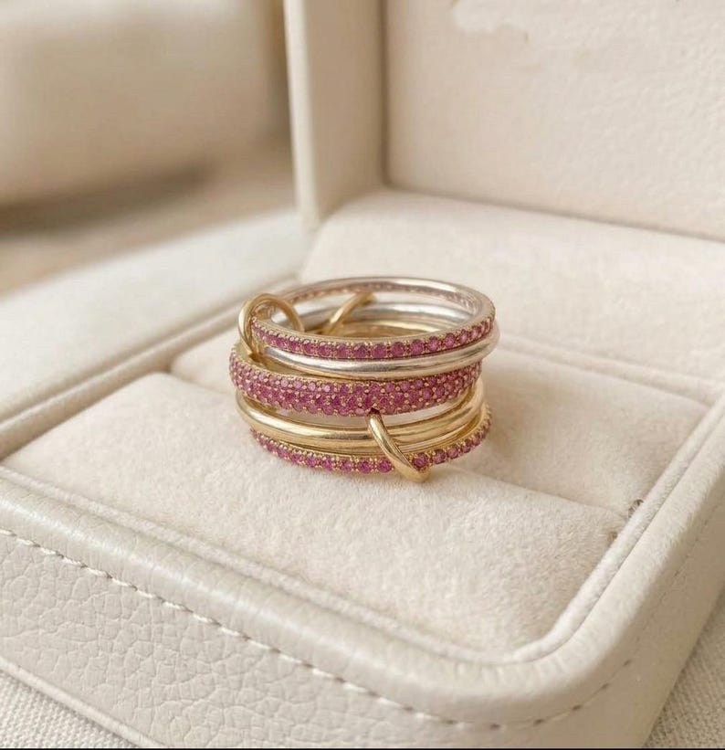 Pu&ograve; includere: Una pila di diversi anelli con fasce alternate in metallo lucido color oro e argento. Molti degli anelli sono incastonati con piccole pietre preziose rosa brillante che coprono l'intera superficie esterna delle fasce circolari.