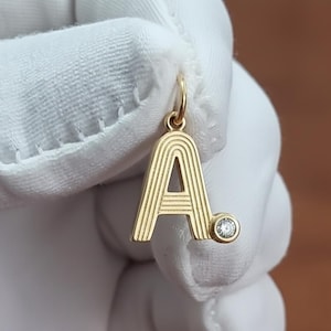 Könnte beinhalten: Goldfarbener Anhänger in Form des Buchstabens "A". Der Anhänger hat ein gestreiftes Design und einen kleinen, runden, klaren Edelstein. Der Anhänger ist an einer kleinen Schlaufe aufgehängt.