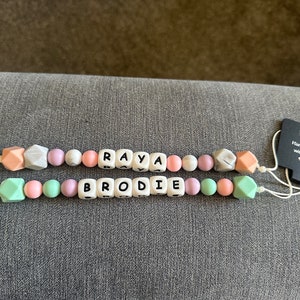 Puede incluir: Un mordedor de bebé de silicona blanco con los nombres "Raya" y "Brodie" escritos en letras negras. El mordedor tiene una variedad de cuentas de colores pastel, incluyendo rosa, verde y morado. El mordedor está unido a dos clips blancos. Una etiqueta negra con el texto "Hecho a mano con amor" está unida al mordedor.