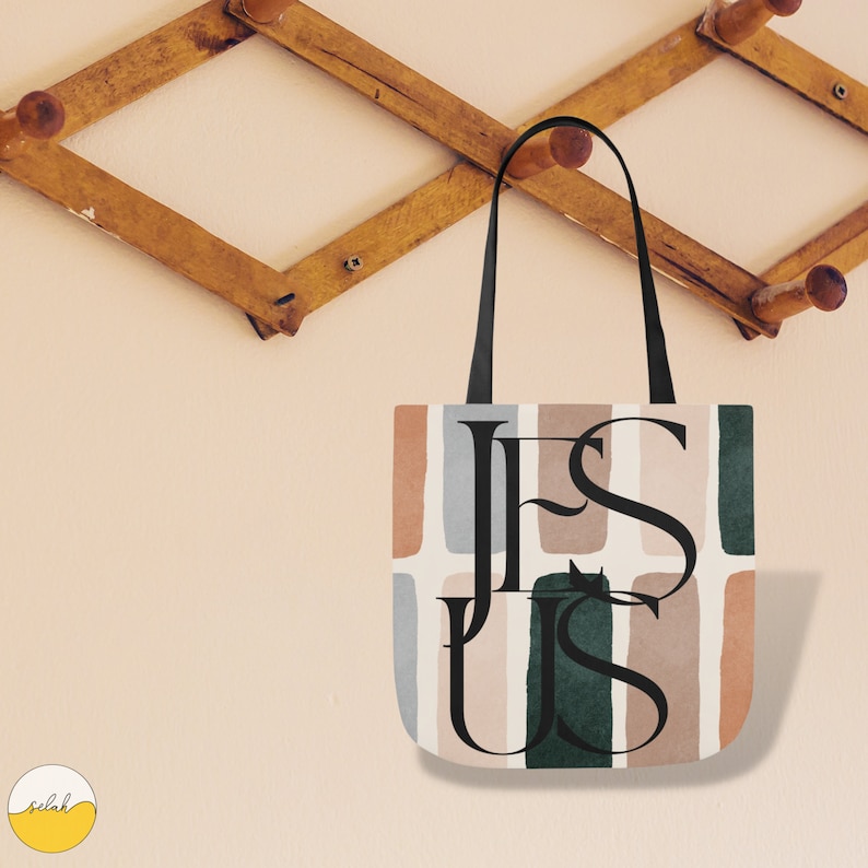 May include: A tote bag with a white background and abstract shapes in shades of brown, green, and blue. The word "JESUS" is printed in black lettering on the bag.