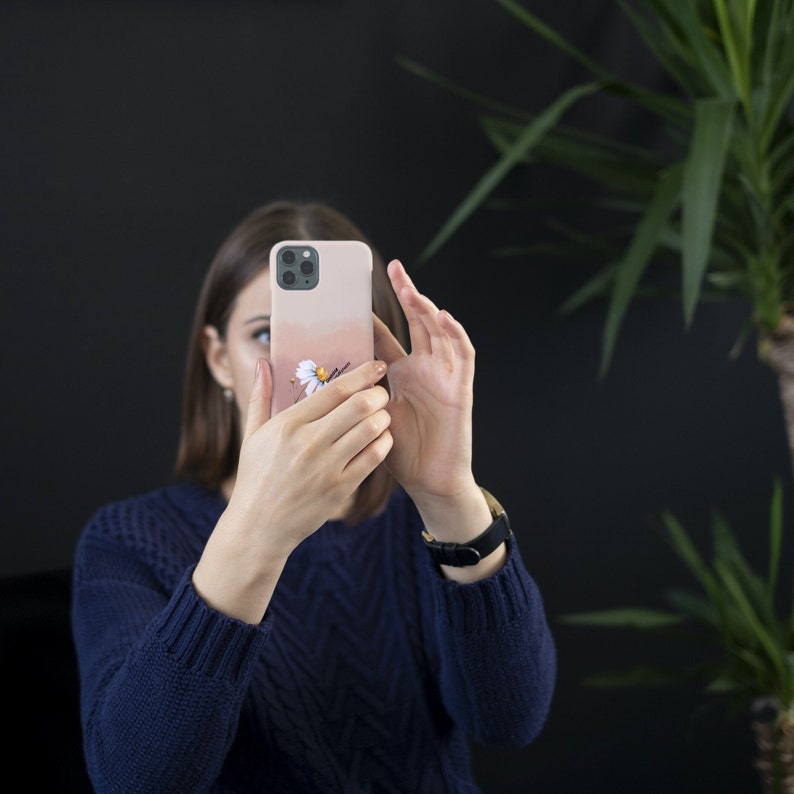 May include: A person is holding a pink and white iPhone case with a floral design. The case has a white daisy with a yellow center and green leaves.