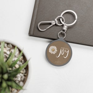Op de afbeelding: Een zilveren sleutelhanger met een ronde hanger met een bruine achtergrond met een witte madeliefje en de tekst "Count it all joy" in witte cursieve letters. De tekst "James 1:2-4" is ook in kleinere witte letters opgenomen.