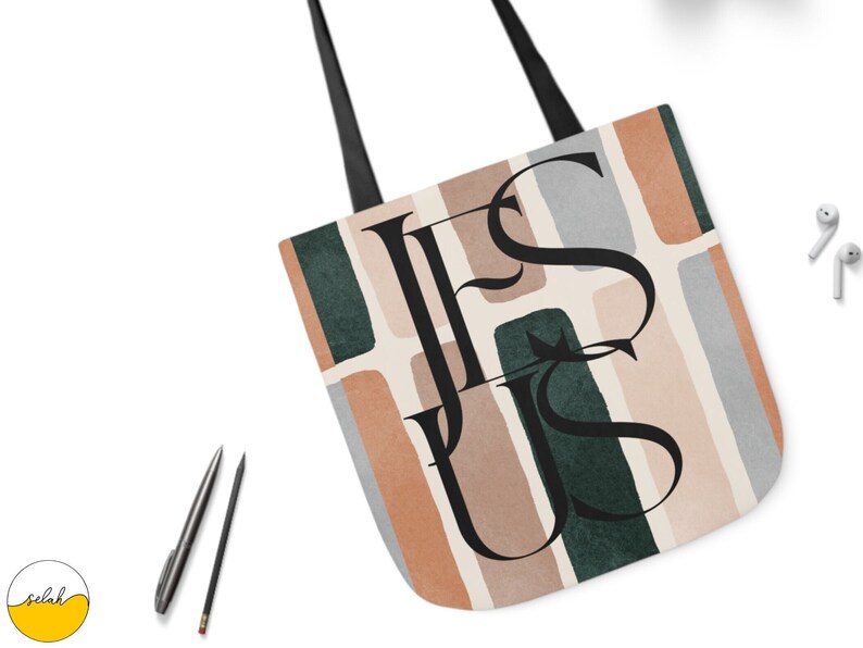 May include: A tote bag with a beige background and abstract shapes in shades of brown, green, and gray. The word "JESUS" is written in black lettering over the shapes.