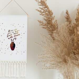 Puede incluir: Tapiz de pared blanco con un jarrón marrón y dorado y el texto "Those who sow in tears shall reap with shouts of joy". El tapiz tiene borlas blancas.