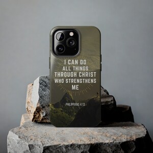 Op de afbeelding: Een groene en bruine telefoonhoes met een berglandschap en de tekst "Ik kan alles door Christus, die mij kracht geeft Filippenzen 4:13".