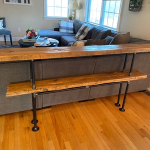Pu&ograve; includere: Un ripiano in legno a due livelli con gambe in tubo metallico nero. Il ripiano &egrave; realizzato in legno riciclato e ha un aspetto rustico. Il ripiano &egrave; posizionato davanti a un divano grigio.
