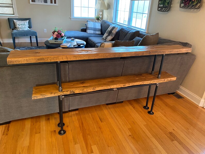 Pu&ograve; includere: Un ripiano in legno a due livelli con gambe in tubo metallico nero. Il ripiano &egrave; realizzato in legno riciclato e ha un aspetto rustico. Il ripiano &egrave; posizionato davanti a un divano grigio.