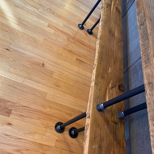 Pu&ograve; includere: Una panca in legno rustico con un piano a bordo vivo e gambe in tubo di metallo nero. La panca &egrave; montata a parete e ha una finitura in legno naturale.