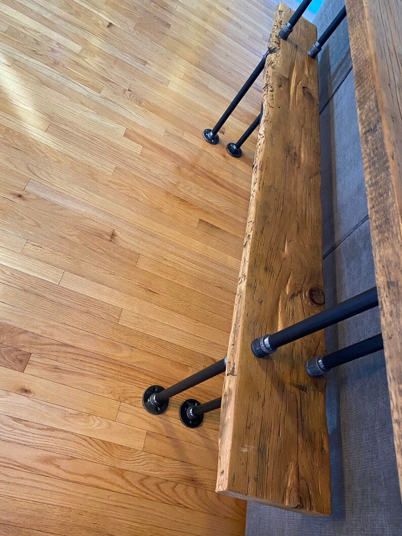 Pu&ograve; includere: Una panca in legno rustico con un piano a bordo vivo e gambe in tubo di metallo nero. La panca &egrave; montata a parete e ha una finitura in legno naturale.