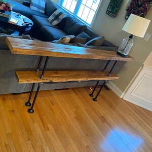 May include: A two-tiered console table made of reclaimed wood with black metal pipe legs. The table is in a living room setting with a couch and a lamp.