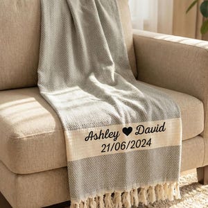 May include: A gray woven throw blanket with a textured pattern and fringe edges. A cream-colored horizontal band features the black printed text "Ashley ♥ David" above the date "21/06/2024" in a simple, dark, sans-serif font style.