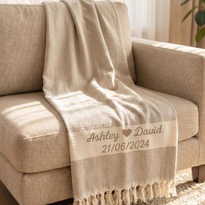 May include: A beige woven throw blanket with a textured herringbone pattern and fringe trim. The blanket features a horizontal cream-coloured band with the embroidered text "Ashley ♥ David" and the date "21/06/2024" in a dark brown script font.