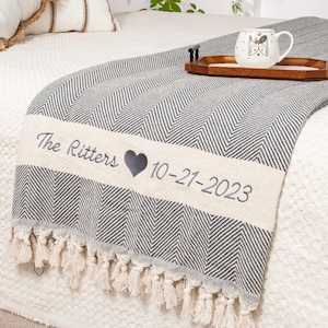 Peut inclure: Une couverture grise et blanche à chevrons avec une bordure crème. La bordure est brodée avec "The Ritters", un cœur et "10-21-2023". La couverture a des franges et est sur un lit avec des oreillers et une tasse.