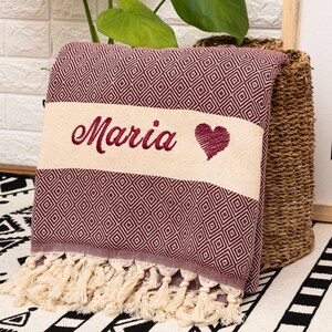 Puede incluir: Una manta plegada de color burdeos y crema con un patr&oacute;n de diamantes y flecos. La manta tiene el nombre "Maria" bordado en burdeos, junto con un s&iacute;mbolo de coraz&oacute;n. La manta es probablemente una manta decorativa o una toalla de playa.
