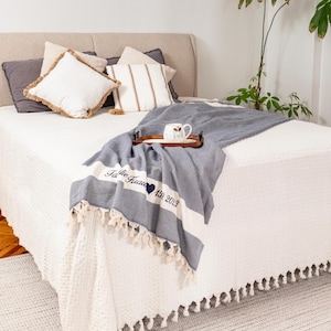 Puede incluir: Una cama con ropa de cama blanca y cojines decorativos. Una manta azul y blanca con borlas est&aacute; sobre la cama. La manta tiene el texto "The Garcia 159. 2023". Una bandeja de madera sostiene una taza.