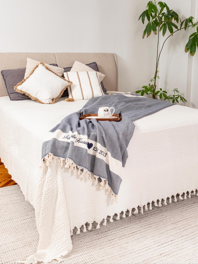 Puede incluir: Una cama con ropa de cama blanca y cojines decorativos. Una manta azul y blanca con borlas est&aacute; sobre la cama. La manta tiene el texto "The Garcia 159. 2023". Una bandeja de madera sostiene una taza.