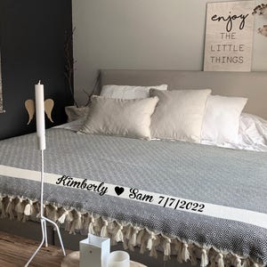 Puede incluir: Una cama con una manta estampada gris y blanca, adornada con una franja blanca y flecos. La cama est&aacute; vestida con varias almohadas blancas y beige. Un letrero de madera dice "enjoy the LITTLE THINGS". Una vela blanca y alas decorativas est&aacute;n a un lado.