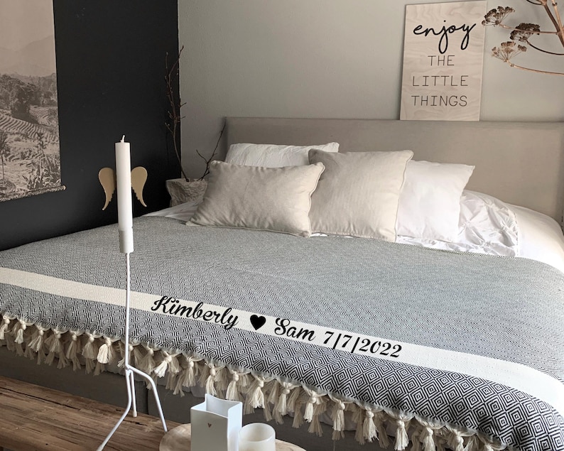 Puede incluir: Una cama con una manta estampada gris y blanca, adornada con una franja blanca y flecos. La cama est&aacute; vestida con varias almohadas blancas y beige. Un letrero de madera dice "enjoy the LITTLE THINGS". Una vela blanca y alas decorativas est&aacute;n a un lado.