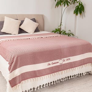 Puede incluir: Una manta a rayas rojas y blancas con un borde con flecos est&aacute; sobre una cama. La manta tiene el texto "The Camerons Est. 2024" en una fuente decorativa. Se ven dos almohadas.