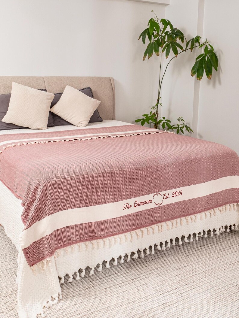 Puede incluir: Una manta a rayas rojas y blancas con un borde con flecos est&aacute; sobre una cama. La manta tiene el texto "The Camerons Est. 2024" en una fuente decorativa. Se ven dos almohadas.