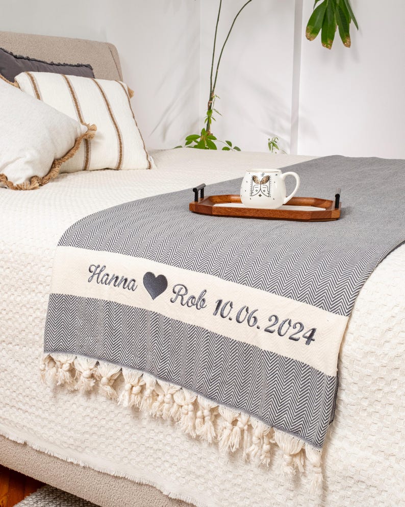 Puede incluir: Una manta gris y blanca con estampado de espiga y borde crema. El borde est&aacute; bordado con los nombres "Hanna" y "Rob", un s&iacute;mbolo de coraz&oacute;n y la fecha "10.06.2024". La manta tiene un ribete de flecos. Una taza y una bandeja encima.