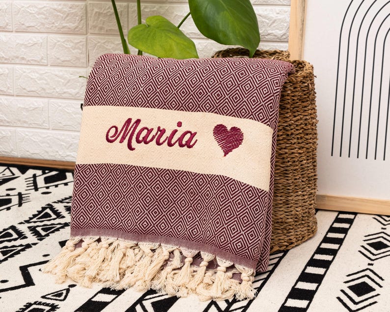 Puede incluir: Una manta plegada de color burdeos y crema con un patr&oacute;n de diamantes y flecos. La manta tiene el nombre "Maria" bordado en burdeos, junto con un s&iacute;mbolo de coraz&oacute;n. La manta es probablemente una manta decorativa o una toalla de playa.