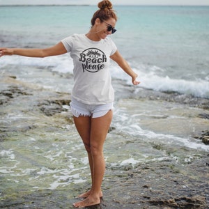 May include: A white t-shirt with a black circle graphic that says "Beach Please" with palm trees inside the circle. The person wearing the shirt is standing on a rocky beach with the ocean in the background.