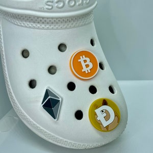 May include: White Crocs shoe with three cryptocurrency-themed charms. The charms include a Bitcoin symbol in orange, an Ethereum symbol in gray and black, and a Dogecoin charm with a dog illustration.