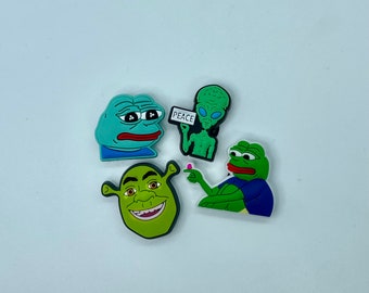 May include: A collection of four colorful cartoon character charms. Includes a green Shrek, a green alien holding a sign that says "PEACE", and two frog-like characters. These charms are likely for shoes or accessories.