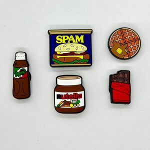 May include: A collection of colorful shoe charms featuring food items. Includes a brown bottle with green and white text, a can of SPAM, a jar of Nutella, a waffle with butter, and a chocolate bar.