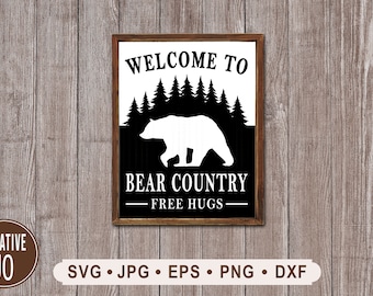 Puede incluir: Un letrero de pared rectangular con un marco de madera marrón oscuro que presenta un gráfico en blanco y negro. El diseño muestra la silueta de un oso frente a un bosque de pinos con el texto WELCOME TO BEAR COUNTRY y FREE HUGS debajo.