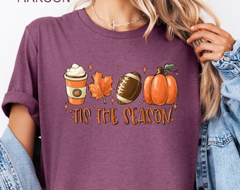 Puede incluir: Una camiseta color granate jaspeado con un estampado gráfico de una taza de café, una hoja de otoño, un balón de fútbol americano y una calabaza. Debajo de los iconos, el texto 'TIS THE SEASON' aparece en una fuente naranja estilizada.