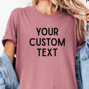 May include: Heather mauve t-shirt with the text "YOUR CUSTOM TEXT" in black. The model is wearing a light blue denim jacket over the shirt. The shirt is a solid color and has rolled-up sleeves.
