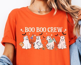 Puede incluir: Una camiseta naranja con un estampado gráfico que incluye el texto "BOO BOO CREW" sobre cuatro fantasmas de dibujos animados vestidos como profesionales médicos, con gorros de enfermera, portapapeles, jeringuilla y botiquín, decorado con murciélagos.