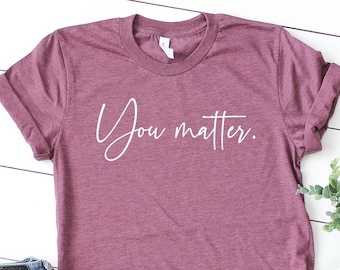 May include: A maroon t-shirt with the text "You matter." in white script lettering. The shirt is laid flat on a white wooden surface with a pair of blue jeans and white sneakers.