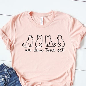 May include: A pink t-shirt with a graphic of three cats and the text "un deux trois cat". The shirt is paired with blue jeans and white sneakers.