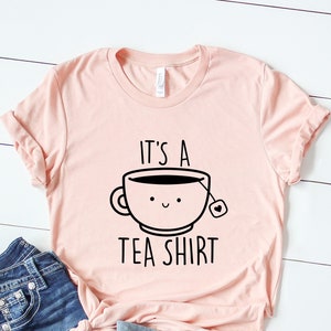 May include: A pink t-shirt with a black graphic of a teacup with a smiling face and the text "It's a Tea Shirt".