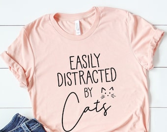 Puede incluir: Una camiseta rosa con el texto "Easily Distracted by Cats" y un pequeño gráfico de cara de gato. La camiseta se combina con jeans azules y zapatillas blancas.