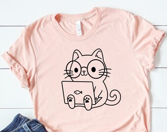 May include: A light pink t-shirt featuring a black and white cartoon cat wearing glasses and holding a laptop with a fish icon. The shirt is paired with blue jeans and white sneakers, arranged on a white wooden surface.