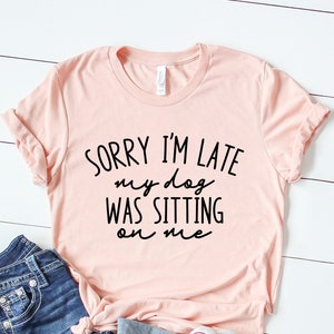 Op de afbeelding: Een roze T-shirt met de tekst "Sorry, ik ben te laat, mijn hond zat op me" in zwarte letters.