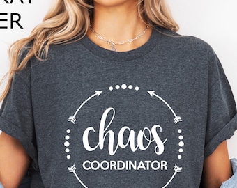 Puede incluir: Camiseta de color gris oscuro jaspeado con la frase "chaos coordinator" en un diseño circular blanco. El texto está rodeado de flechas y puntos. La camiseta es de cuello redondo y tiene mangas enrolladas.