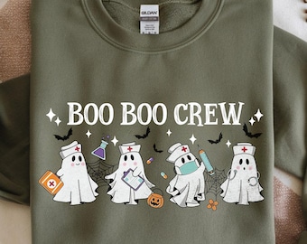Puede incluir: Sudadera verde oliva con el texto "BOO BOO CREW" e ilustraciones de personajes fantasma vestidos de enfermeras, con murciélagos y estrellas. El diseño incluye una calabaza, medicinas y una telaraña.