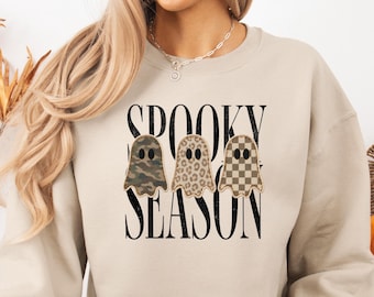 Puede incluir: Una sudadera beige de cuello redondo con el texto negro "SPOOKY SEASON" y tres ilustraciones de fantasmas. Los fantasmas tienen estampados de camuflaje, leopardo y cuadros, cada uno con dos ojos ovalados negros y una boca pequeña.