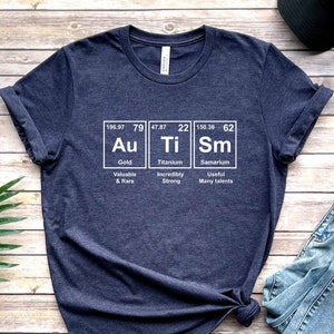 May include: A navy blue t-shirt with a white graphic of the periodic table elements Au, Ti, and Sm. The graphic includes the atomic number, atomic weight, and name of each element. The text below each element describes its properties: Gold - Valuable & Rare, Titanium - Incredibly Strong, Samarium - Useful Many talents.
