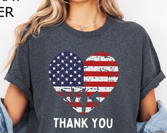 Puede incluir: Camiseta gris oscuro jaspeado con un diseño de bandera americana en forma de corazón. El corazón está dividido en dos mitades, una con estrellas y la otra con rayas. Las palabras "THANK YOU" están impresas debajo.