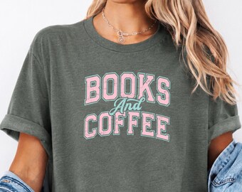 May include: A heather military green short-sleeve t-shirt with a crew neckline. The front features the text "BOOKS And COFFEE" in a distressed, collegiate-style font with pink lettering and a light blue script for the word "And".