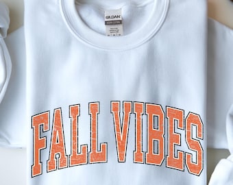 Puede incluir: Sudadera blanca con el texto "FALL VIBES" en letras naranja. La sudadera tiene cuello redondo y mangas largas con puños enrollados. Una pequeña calabaza y hojas de otoño están en el fondo.