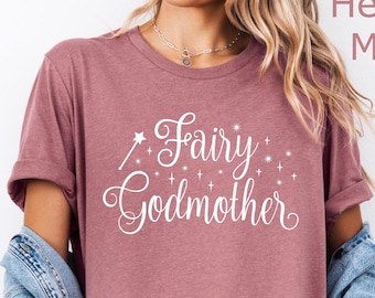 Puede incluir: Camiseta color malva con la frase "Fairy Godmother" en escritura blanca. La camiseta tiene cuello redondo y mangas cortas. La modelo lleva una chaqueta vaquera y pantalones cortos.