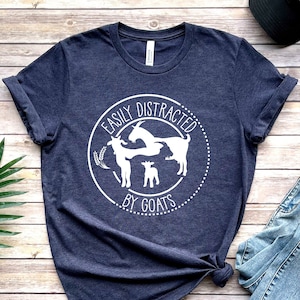 May include: A navy blue t-shirt with a white circle design that says "Easily Distracted by Goats". The design features three goats, two adults and a baby goat.